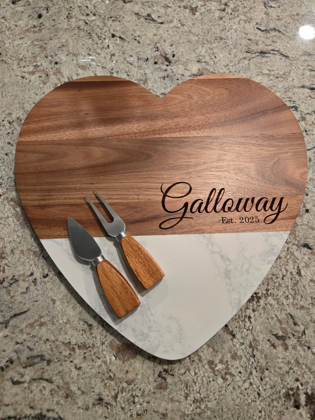 Heart-shaped wooden serving board with a white marble lower section, engraved with the name 'Galloway Est. 2025', and shown with two small wooden-handled serving utensils on a granite countertop.