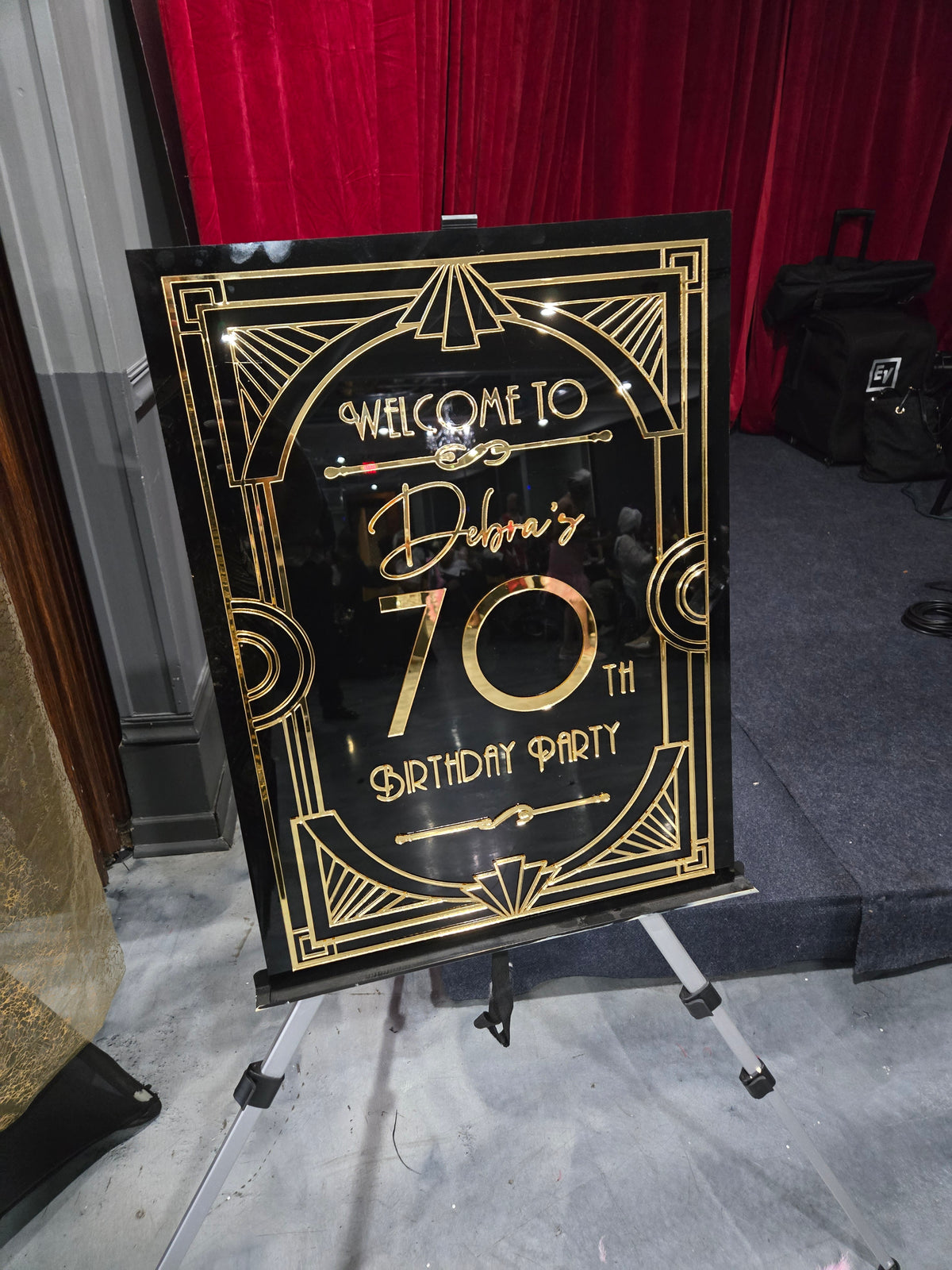 Decorative birthday sign with gold text on a black background, placed in front of a red drape and stage
