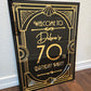 Decorative birthday party sign with gold text on a black background, leaning against a wall.